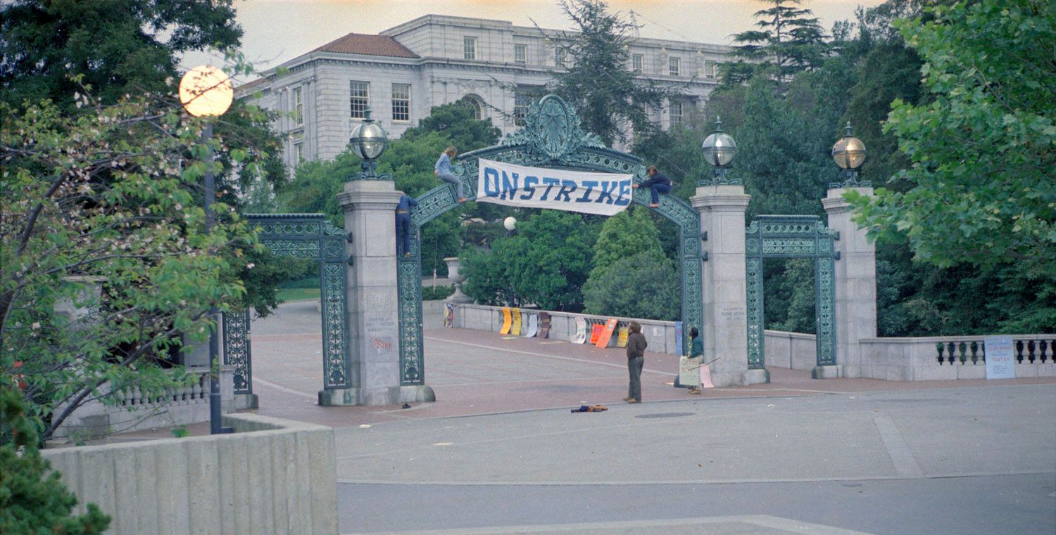 Several students around a deserted Sather Gate in the evening, raising a banner that says On Strike